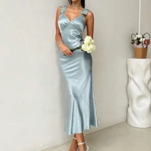 Ljusblå satinklänning med blommiga axelband - Helt ny med prislapp!!!      Superfin ljusblå långklänning i satin med v-ringning och smala axelband dekorerade med blommiga spetsdetaljer. Klänningen har en figurnära passform och faller snyggt längs kroppen. Perfekt för sommarens speciella tillfällen.   