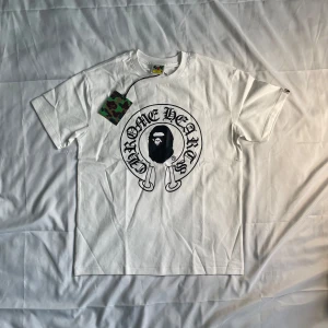 Chrome Hearts X Bape T skjorta - Helt ny vit t-shirt – Storlek M – Chrome Hearts x BAPE Inspired  🆕 Skick: Helt ny, aldrig använd 📏 Storlek: M (Medium) 🎨 Färg: Vit 👕 Design: Djärv grafik framtill med en unik logotyp inspirerad av Chrome Hearts och A Bathing Ape (BAPE). Iögonfallande svart tryck på en skarp vit tröja.  🔖 Detaljer: Levereras med original kamouflage-etikett.  🔥 Perfekt för streetwear-älskare och samlare. Högkvalitativ bomull med en ikonisk design. Köp den innan den är borta!  📦 Skickas snabbt och omsorgsfullt!