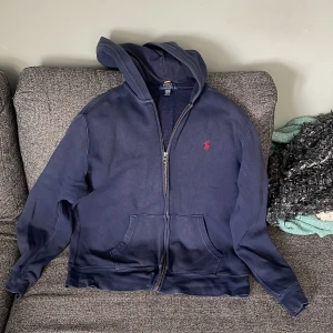 Marinblå hoodie från Polo Ralph Lauren XL (barn)  - Säljer en marinblå hoodie från Polo Ralph Lauren med dragkedja och huva. Klassisk röd logga broderad på bröstet och två fickor framtill. Perfekt för dig som gillar en avslappnad och stilren look.