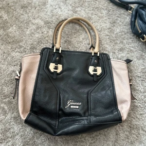 Svart och beige handväska från Guess - Snygg handväska från Guess i svart och beige skinn med stilrena detaljer i guld. Väskan har två handtag, dragkedja upptill och flera innerfack med logomönstrat foder. Perfekt storlek för allt du behöver ha med dig och en klassisk look.