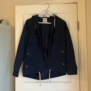 Mörkblå parkajacka från ONLY - Jacka i mörkblått från ONLY med huva, dragkedja och snörning nertill. Jackan har två stora bröstfickor med lock och två sidofickor med snygga detaljer. Perfekt för dig som gillar en clean och avslappnad stil.