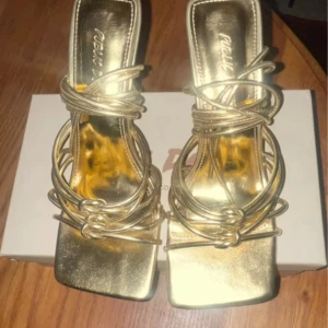 Guldiga sandaler med klack och snörning - Snygga guldiga sandaler från Public Desire med fyrkantig tå, höga klackar och trendiga snörningsdetaljer runt foten. Skorna har metallicfinish och öppen tå, perfekta för att sticka ut. Passar dig som gillar statement-accessoarer och vill ha något extra till outfiten.Mycket fint skick se bilder lite intryckt på ena klacken. Annars använda bara en kväll.  Stl 36