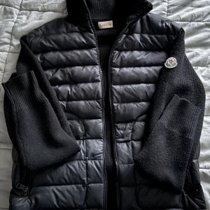 Moncler cardigan  - Snygg svart dunväst från Moncler med quiltad framsida och stickade ärmar och rygg. Västen har hög krage, två dragkedjefickor och Moncler-logga på ärmen. Perfekt för lager-på-lager och riktigt stilren look.