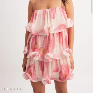 Rosa blommig volangklänning - Superfin kortklänning med stora rosa blommor och volangdetaljer. Klänningen har tunna axelband, är ärmlös och har ett luftigt, plisserat tyg som ger en riktigt somrig vibe. Klänningen är helt ny med prislapp kvar. Nypris var 699kr