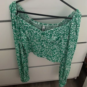 Grön blommig blus - Supersöt grön blommig blus från New yorker i XS. Modellen är croppad med långa ärmar och v-ringning. Blusen har små knappar framtill och elastisk resår vid axlarna för en offshoulder-look, finns även resår i ryggen. Perfekt för vår och sommar!