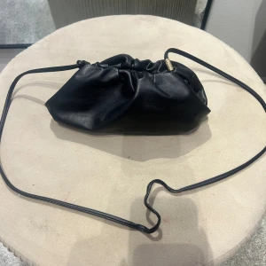 Svart axelväska från BOW19 Details - Stilren svart axelväska i mjukt skinn från BOW19 Details. Väskan har en unik rynkad design med smal axelrem och guldfärgade metalldetaljer. Perfekt storlek för det viktigaste och enkel att bära crossbody eller på axeln.