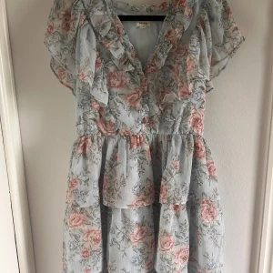 Blommig volangklänning från Newbie - Supersöt kort blommig klänning från Newbie i ljusblått med rosa och gröna blommor. Klänningen har volanger längs ärmar och halsringning, korta ärmar och en luftig passform. Perfekt för sommarens alla tillfällen!