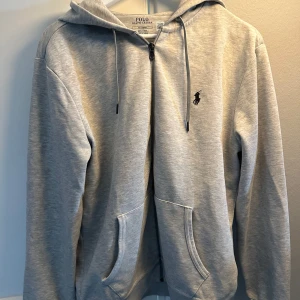 Grå hoodie från Polo Ralph Lauren - Snygg ljusgrå hoodie från Polo Ralph Lauren med dragkedja framtill, huva med snörning och klassisk broderad logga på bröstet. Tröjan har två fickor och är tillverkad i mjuk bomullsmix. Perfekt för en chill och stilren look.