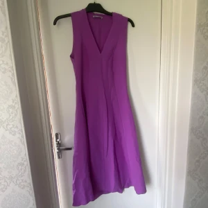 Lila ärmlös klänning med v-ringning - Snygg lila klänning med v-ringad hals och ärmlös design. Klänningen har en lätt utställd kjol och stängs med dragkedja i sidan. Perfekt för dig som vill sticka ut med en färgstark look. Materialet känns mjukt och följsamt.