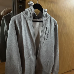 Grå hoodie från Sail Racing XL - Snygg grå hoodie från Sail Racing i storlek XL. Tröjan har dragkedja, huva och en bröstficka med zip. Insidan är mjukt fodrad med fleece som gör den mycket skön. Perfekt till casual tillfällen eller bara att ha hemma. Den är nästintill aldrig använd och är i mycket bra skick. Ordinarie pris 1100.