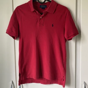 Röd pikétröja från Polo Ralph Lauren - Röd Polo Ralph Lauren Piké med korta ärmar och krage. Tröjan har två knappar och broderade loggan i mörkblått på bröstet. Tillverkad i bomull och det finns små hål vid vänstra bröstet (se sista bilden) men inget som man märker. 10 - 12 år. Originalpris : 800-1000 kr. Pris kan diskuteras