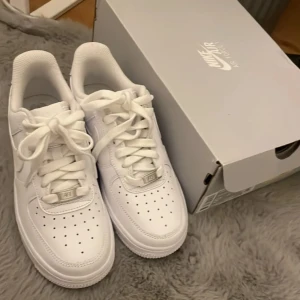 Nike Air Force 1  - Klassiska Nike Air Force 1 sneakers i helvitt skinn med perforerad tå, rund tå och platt sula. Snygg och tidlös modell med snörning och diskret Nike-logga på sidan. Perfekta för dig som vill ha en clean och fräsch look.