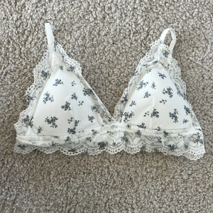 Blommig spets bralette från H&M - Supersöt vit bralette från H&M med små blå blommor och romantisk spetskant runtom. Triangelmodell med justerbara axelband och mjukt ribbat tyg.  Är i storlek 80C/M
