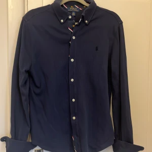 Mörkblå skjorta från Polo Ralph Lauren - Klassisk mörkblå skjorta från Polo Ralph Lauren med button down-krage och vita knappar. Skjortan har lång ärm och diskret broderad logga på bröstet. Tillverkad i mjuk bomull som känns skön mot huden. Perfekt för en stilren look.