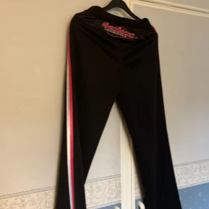 Svarta träningsbyxor Lanshuang Made in China - Svarta träningsbyxor från Lanshuang med broderad logga både fram och bak. Byxorna har vita och rosa ränder längs sidorna och är tillverkade i ett glansigt syntetmaterial. Perfekta för dig som gillar sportig stil och vill sticka ut med coola detaljer. Storlek L 165/69 Kina. men är i svensk storlek xs/s tänker jag. Beninnerlängd 74 cm. Benvidd nedtill 21-22 cm.Midja mätt runt tyget upptill 64. cm. Två sidfickor utan dragkedja. Första bild byxans baksida. Bild 2 framsida. Bild 3 ficka. 