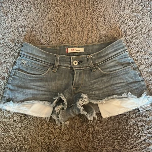 Levi's jeansshorts  - Snygga blå jeansshorts från Levi's med fransig och sliten kant. Klassisk femficksmodell, låg midja och gul kontrastsöm. Perfekta för sommaren och ger en cool, avslappnad vibe. Levi's logga på bakfickan och läderpatch bak i midjan. Köpta i barcelona vintage väldigt svåra att få tag på.