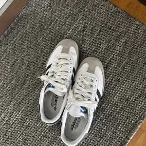 Adidas Samba - Klassiska Adidas Samba sneakers i vitt skinn med grå mockadetaljer och svarta ränder på sidorna. Skorna har snörning och en platt sula, perfekt för en sportig och stilren look. Ikonisk blå Adidas-logga på plösen och svart detalj på hälen. Nypris 1400
