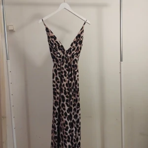 Leopardmönstrad långklänning med v-ringning - Snygg långklänning med leopardmönster och djup v-ringning. Klänningen har tunna axelband och ett lätt, flytande tyg som ger en cool och trendig look. Perfekt för dig som vill sticka ut med ett djuriskt mönster och en feminin siluett.