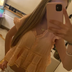 Persikofärgad volangblus från H&M - Supersöt persikofärgad blus från H&M med volanger, v-ringning och knappar framtill. Blusen har korta ärmar och är tillverkad i ett lätt, transparent material med fina detaljer och rynkade kanter. Perfekt för en somrig och romantisk look!