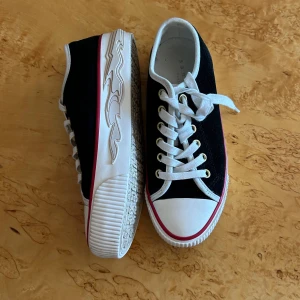 Svarta sneakers från Sandro Paris - Coola svarta sneakers från Sandro Paris med vita detaljer, vit tå och sula med röd rand. Snygga guldfärgade öljetter och vita snören. Sidan av sulan har ett vågigt mönster som ger extra edge. Perfekta för dig som gillar stilrena men unika skor.
