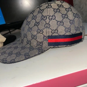 Gucci keps med GG-mönster och röd/blå detalj - Snygg keps från Gucci i beige med mörkblått GG-monogram över hela kepsen. På sidan finns en ikonisk röd och blå rand. Kepsen är tillverkad i bomull och har klassisk böjd skärm. Perfekt för dig som vill sticka ut med en lyxig accessoar.