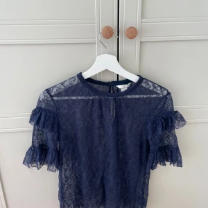 Marinblå spetsblus från H&M - Säljer en marinblå blus från H&M i genombruten spets med volangdetaljer på ärmarna. Blusen har rund halsringning, korta ärmar och knäppning bak i nacken🥰