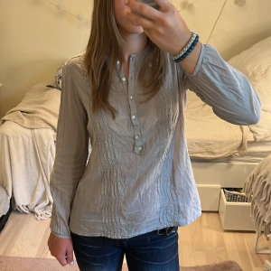 Fin blus - Ljusgrå långärmad blus med knappar framtill (går att knäppa upp hela vägen). Tröjan har sydda detaljer längs bröstet och knapp i ärmens slut.
