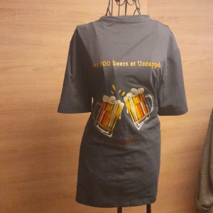 Grå t-shirt med ölglas-tryck, L - Grå t-shirt i 100% polyester med coolt ölglas-tryck och texten 'Over 100 beers at Untappd' på bröstet. Klassisk passform med korta ärmar och rund hals. Perfekt för dig som gillar unika och roliga motiv.