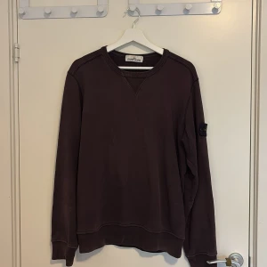 Mörkbrun Stone Island sweatshirt - Säljer en mörkbrun sweatshirt från Stone Island med klassisk rund hals och ribbade muddar. Tröjan har den ikoniska loggan på vänster ärm och är tillverkad i mjuk bomull. Perfekt för dig som gillar streetwear och snygga detaljer.