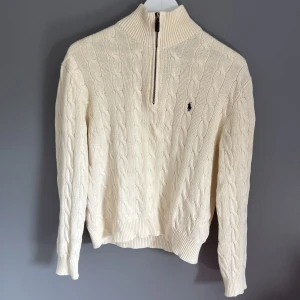 Vit kabelstickad tröja Polo Ralph Lauren - Kabelstickad vit tröja från Polo Ralph Lauren med half zip och hög krage. Klassisk design med broderad logga på bröstet och ribbade muddar. Perfekt för lager-på-lager och snygg till jeans eller chinos. 