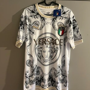 Italien Versace x Puma fotbollströja - Snygg vit fotbollströja med svart och guld barockmönster, Versace-logga i guld framtill och Italiens landslagsemblem på bröstet. Guldiga detaljer vid hals och ärmslut. Tillverkad i lätt och glansigt material, perfekt för dig som vill sticka ut på planen.