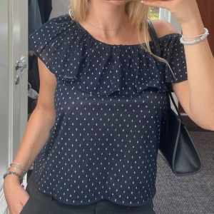 Mörkblå offshoulder blus med volang - Supersöt mörkblå blus med offshoulder och volang runt halsen. Små vita mönster ger en fräsch känsla och toppen är luftig och perfekt för varma dagar. Kan bäras på olika sätt. Både över och under armarna.