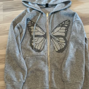 Grå hoodie med fjärilstryck - Grå hoodie med dragkedja och snörning i huvan. Stort svartvitt fjärilstryck på framsidan som ger en cool och unik look. Perfekt för dig som gillar streetstyle och vill sticka ut lite extra.
