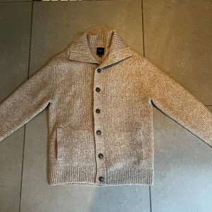 Beige stickad cardigan jacka från Next - Stickad cardigan jacka i beige från Next med grov ribbad struktur, stora bruna knappar och två fickor framtill. Cardiganen har hög krage och långärmad passform, perfekt för lager-på-lager under kyliga dagar. Stor i storleken