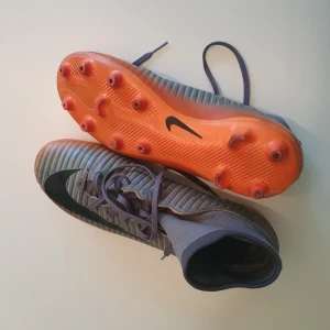 Nike Mercurial fotbollsskor grå/orange (sulor ingår inte) - Säljer ett par Nike Mercurial fotbollsskor med grå ovandel och svarta detaljer. Skorna har högt skaft i stretchigt material, snörning och orange sula med fasta dobbar. Perfekta för dig som vill ha grymt grepp och snygg stil på planen.