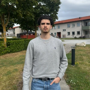 Grå sweatshirt från Polo Ralph Lauren - Klassisk grå sweatshirt från Polo Ralph Lauren med rund halsringning och broderad marinblå logga på bröstet. Tröjan har ribbade muddar vid ärmslut och midja, och är tillverkad i mjuk bomullsmix. Perfekt för en clean och avslappnad stil.
