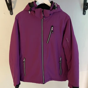 Damjacka ifrån Tuxer i stl L - Snygg lila jacka från Tuxer med svart och vit dragkedja, justerbar huva och flera fickor med dragkedja. Jackan har långa ärmar och reflekterande detaljer längs ryggen och ärmarna. Perfekt för blåsiga dagar och outdoor-aktiviteter.