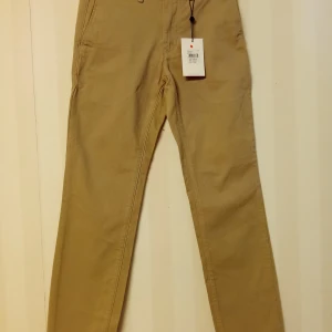 Beige chinos från These Glory Days - Snygga beige chinos från These Glory Days i klassisk rak modell. Byxorna har dragkedjegylf, bälteshällor och två bakfickor, varav en med knapp. Tillverkade i bomull för en skön och stilren känsla.