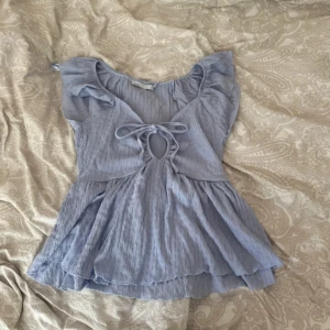 Ljusblå blus med volang och knyt - Supersöt ljusblå blus med volangdetaljer och knyt framtill. Toppen har korta ärmar och en flowy, luftig passform. Perfekt för dig som gillar romantisk och feminin stil. Materialet är tunt och ger en lätt känsla.