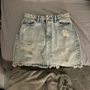 Ljusblå jeanskjol från FB Sister/newyorker - Säljer en ljusblå, (sliten)jeanskjol från FB Sister i storlek M. Kjolen har råa kanter, flera slitningar och fransar för en cool, avslappnad vibe. Klassisk femficksmodell med bälteshällor och dragkedja framtill. Perfekt för dig som gillar sommaren.