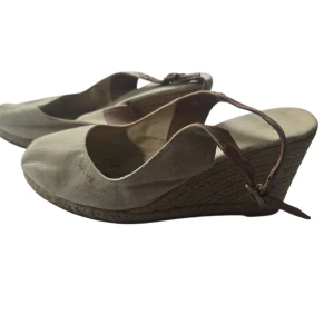 Beige kilklackar från Gant - Snygga beige kilklackar från Gant med ovandel i canvas och bruna läderdetaljer. Skorna har öppen häl med justerbart spänne och flätad sula i espadrillestil. Perfekta för en somrig look med avslappnad känsla.