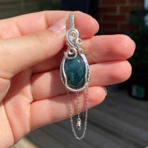 Silverberlock grön sten (mossagat) - Unik berlock med en oval, mörkgrön sten infattad i detaljerad silvertråd. Hängsmycket har snygga snirkliga mönster och två tunna kedjor som dekoration. Perfekt statement piece för dig som gillar boho eller magisk vibe.