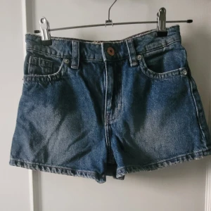 Blå jeansshorts från H&M Divided - Klassiska blå jeansshorts från H&M Divided med normal passform i mid rise. De har fem fickor, bälteshällor och snygga kontrastsömmar. Perfekta för sommaren och lätta att matcha med allt. Stängs med knapp och dragkedja framtill. Passar XS