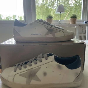 Golden Goose Superstar sneakers vit/grå - Golden Goose Superstar sneakers i vitt skinn med grå stjärndetalj på sidan, grå snörning och mörkblå häl. Snyggt slitna detaljer och platt sula ger en cool, avslappnad vibe. Kommer med originalpåse och pass. Perfekta för dig som gillar streetstyle.