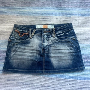 Jeanskjol från Miss Chic med detaljer - Snygg kort jeanskjol från Miss Chic i tvättad blå denim. Kjolen har broderade bakfickor med glittriga paljetter och stenar i gult och brunt. På lappen står det att kjolen är i storlek 36 men jag skulle säga att den är i storlek 34. Kjolen har inga slitningar förutom några saknade paljetter, annars är den använd varsamt!💕