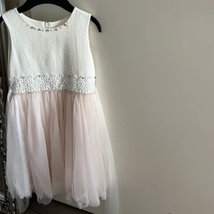 Vit och ljusrosa klänning med pärlor - Supersöt ärmlös klänning med vit strukturerad överdel, dekorerad med pärlor och strass vid halsen samt broderad spets i midjan. Kjolen är i flera lager ljusrosa tyll som ger en fluffig look. Dragkedja bak för enkel påklädning.