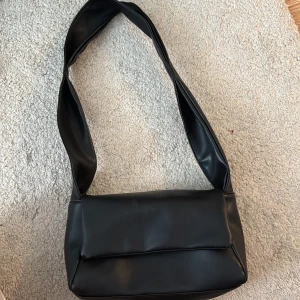 Svart axelväska i skinnimitation - Snygg svart axelväska med bred rem och stilren design. Väskan har ett lock med magnetknappar och är gjord i mjuk skinnimitation. Perfekt storlek för det viktigaste och passar till många olika outfits.