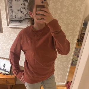 Rostbrun sweatshirt från Levi's - Säljer en orange/brun sweatshirt från Levi's med klassisk rund hals och liten röd logotyp på bröstet. Tröjan är i mjuk bomull och har långa ärmar samt ribbade muddar. Perfekt för chill dagar och enkel att matcha med jeans.