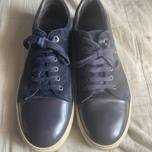 Mörkblå sneakers från Lanvin i mocka - Snygga mörkblå sneakers från Lanvin med ovandel i mocka och tåparti i blankt skinn. Skorna har vita sulor, mörkblå snörning och en detalj i vinrött bak. Perfekta för dig som gillar stilrena och exklusiva sneakers.