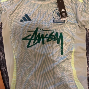 Mexico x Stüssy Adidas fotbollströja S - Unik Mexico landslagströja i samarbete med Stüssy och Adidas. Tröjan är ljusblå med geometriskt mönster, neongula detaljer och tryckt Stüssy-logga på bröstet. Broderat Mexiko-märke och Adidas-logga. Tillverkad i lätt, ventilerande material.
